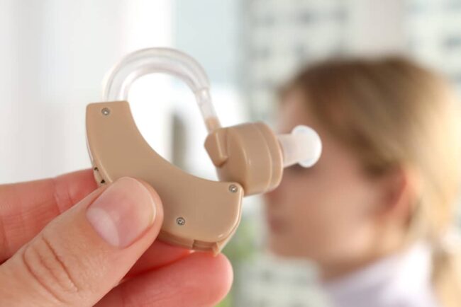 Concept of health care with hearing aid, close up a large beige hearing aid in the foreground in front of a blonde woman's head that is out of focus in the background