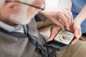 an elderly man with white hair taking a hearing aid out of a box being held by a woman out of frame.
