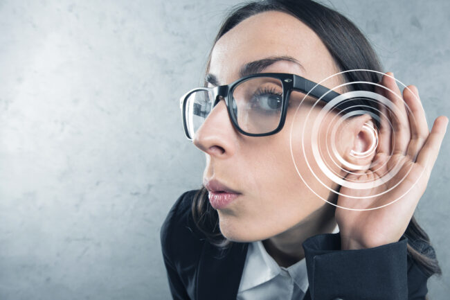 A woman in a black suit wearing glasses holding her hand up to her ear.