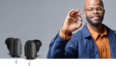 A black man in a blue jacket and orange shirt holding one of the Signia Insio Charge&Go CIC IX to show its small size with a larger image of the hearing aids to provide detail.